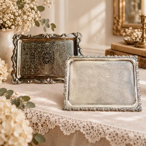 May include: Two rectangular silver-toned trays with ornate, raised floral and scrollwork borders. One tray features a detailed, embossed filigree pattern across its centre, while the other has a smooth, reflective surface with a plain interior.