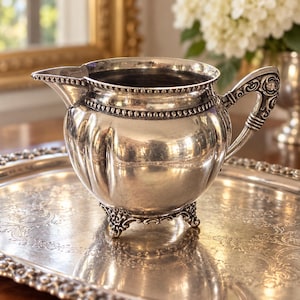 May include: A silver-plated creamer pitcher featuring a rounded, ribbed body, a beaded rim, and an ornate, scrolled handle. The pitcher stands on three decorative, detailed feet and includes a small pouring spout, reflecting a polished metallic finish.