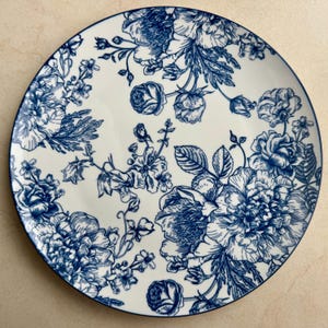 Puede incluir: Un plato redondo de cer&aacute;mica con base blanca y un detallado patr&oacute;n floral en azul oscuro. El dise&ntilde;o consta de varias flores en floraci&oacute;n, capullos y tallos con hojas dispuestos por toda la superficie, rematado con un fino borde azul oscuro.