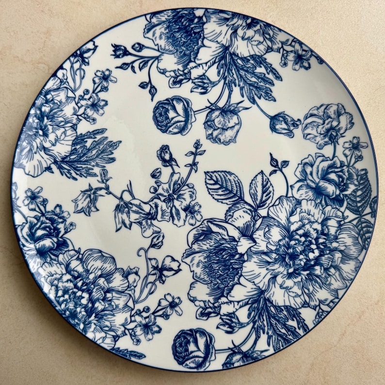 Puede incluir: Un plato redondo de cer&aacute;mica con base blanca y un detallado patr&oacute;n floral en azul oscuro. El dise&ntilde;o consta de varias flores en floraci&oacute;n, capullos y tallos con hojas dispuestos por toda la superficie, rematado con un fino borde azul oscuro.