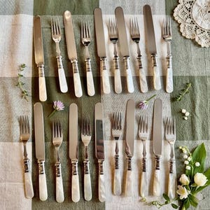 May include: A collection of vintage silverware, including forks and knives, arranged on a green and white checkered tablecloth. The silverware features mother-of-pearl handles and a silver blade. The arrangement includes sprigs of flowers and lace.
