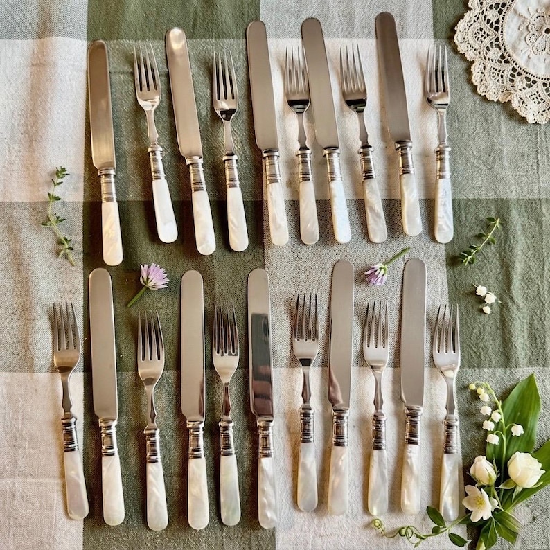 May include: A collection of vintage silverware, including forks and knives, arranged on a green and white checkered tablecloth. The silverware features mother-of-pearl handles and a silver blade. The arrangement includes sprigs of flowers and lace.