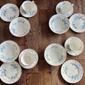 May include: A collection of white porcelain dishware with a delicate blue floral pattern and gold trim. The set includes teacups, saucers, bowls, and plates, arranged on a wooden surface. The plates have a ridged edge.