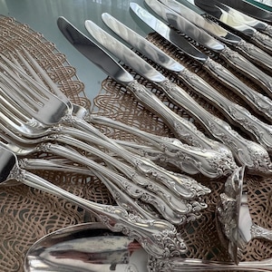May include: An assortment of ornate silver-colored flatware, including forks, knives, and spoons. The handles feature a detailed floral design. The silverware is arranged on a decorative, woven placemat.