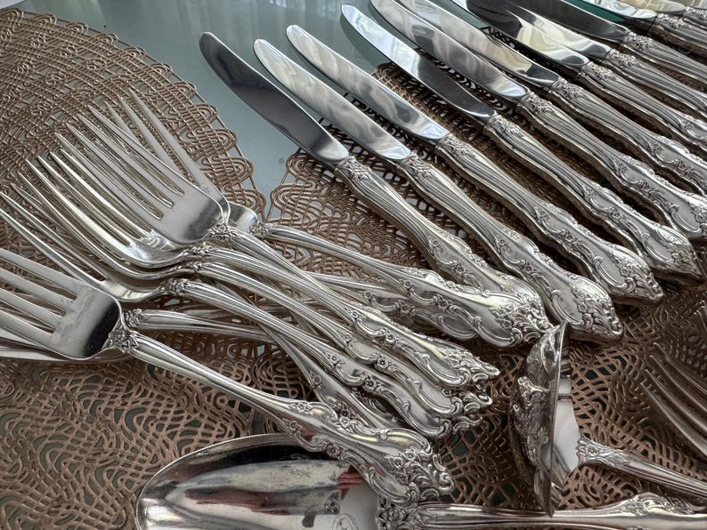 May include: An assortment of ornate silver-colored flatware, including forks, knives, and spoons. The handles feature a detailed floral design. The silverware is arranged on a decorative, woven placemat.