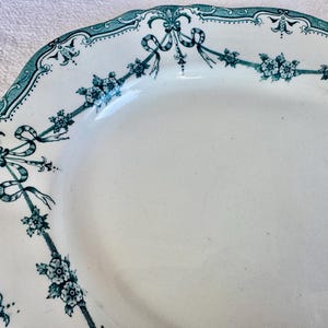 May include: An oval ceramic platter with a scalloped edge, featuring a white base and a teal-coloured decorative border. The border displays a repeating pattern of floral clusters connected by draped ribbons and intricate scrollwork motifs.