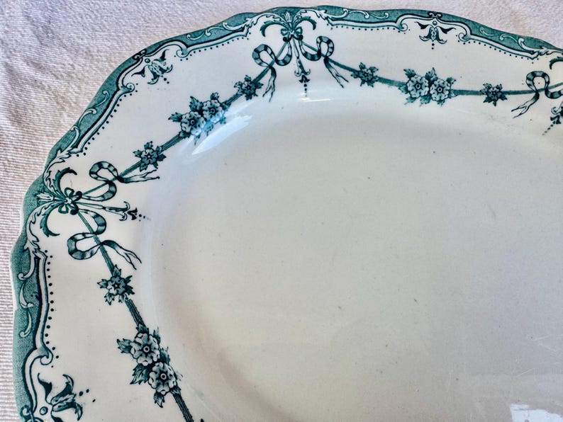 May include: An oval ceramic platter with a scalloped edge, featuring a white base and a teal-coloured decorative border. The border displays a repeating pattern of floral clusters connected by draped ribbons and intricate scrollwork motifs.