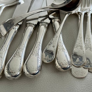 May include: Close-up of a collection of antique silverware. The set includes spoons, forks, and knives with ornate floral engravings on the handles. The silverware is made of a shiny, reflective metal, likely silver.