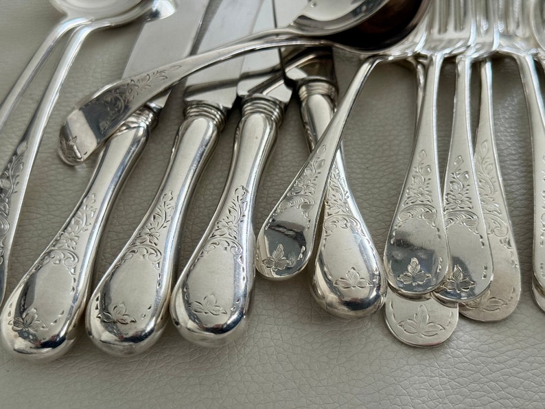 May include: Close-up of a collection of antique silverware. The set includes spoons, forks, and knives with ornate floral engravings on the handles. The silverware is made of a shiny, reflective metal, likely silver.