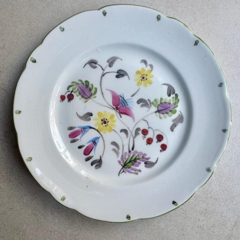 May include: A white ceramic plate with a scalloped edge featuring a hand-painted floral motif in the center. The design includes yellow, pink, and purple flowers with green and gray leaves and small red berries. Tiny green dots accent the rim.
