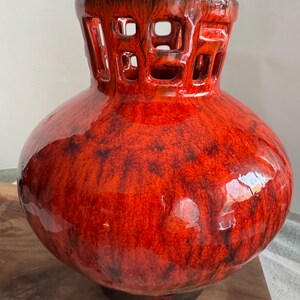 Puede incluir: Un jarr&oacute;n grande de cer&aacute;mica roja brillante con un cuerpo bulboso y un cuello cil&iacute;ndrico con un dise&ntilde;o geom&eacute;trico calado. El jarr&oacute;n tiene una base marr&oacute;n oscuro y una superficie texturizada y moteada, creando un contraste llamativo.