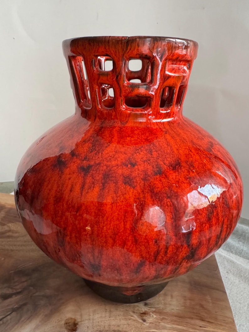 Puede incluir: Un jarr&oacute;n grande de cer&aacute;mica roja brillante con un cuerpo bulboso y un cuello cil&iacute;ndrico con un dise&ntilde;o geom&eacute;trico calado. El jarr&oacute;n tiene una base marr&oacute;n oscuro y una superficie texturizada y moteada, creando un contraste llamativo.
