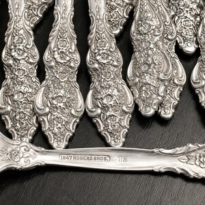 May include: A collection of silver-plated flatware featuring an intricate floral embossed pattern on the handles. The horizontal piece displays the stamped text 1847 ROGERS BROS. and IS. Each handle showcases detailed blossoms and scrolling metalwork.