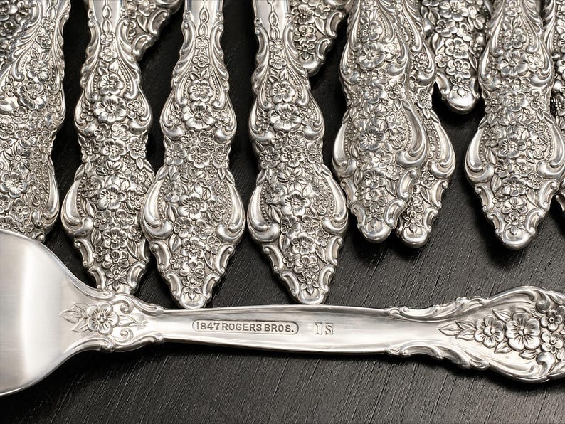 May include: A collection of silver-plated flatware featuring an intricate floral embossed pattern on the handles. The horizontal piece displays the stamped text 1847 ROGERS BROS. and IS. Each handle showcases detailed blossoms and scrolling metalwork.