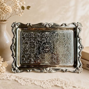 May include: A rectangular silver-toned metal tray featuring an ornate, raised border with intricate scrollwork and a central panel decorated with a detailed, embossed pattern of swirling vines and foliage across the entire flat surface.