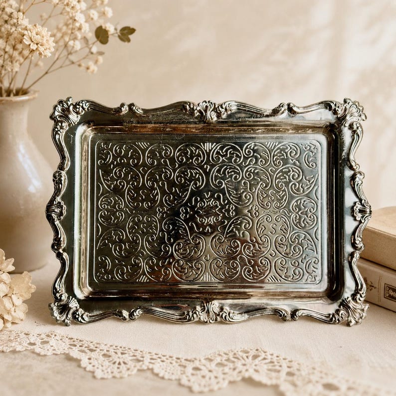Puede incluir: Una bandeja rectangular de metal plateado con un borde elevado y ornamentado con volutas intrincadas y un panel central decorado con un patr&oacute;n en relieve detallado y sim&eacute;trico de enredaderas y follaje.