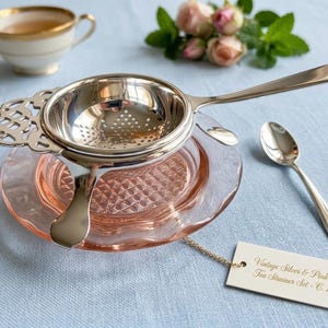 Può includere: Un set di colino da tè vintage con un colino d'argento, un piattino di vetro rosa e un cucchiaino. Il set include un'etichetta che recita "Vintage Silver & Pink Glass Tea Strainer Set - c. 1920s". Una tazza da tè e fiori sono sullo sfondo.