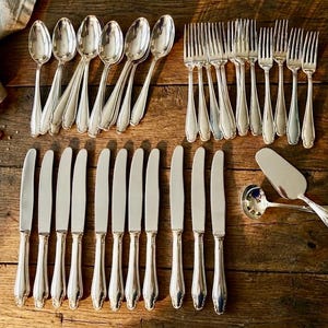 Op de afbeelding: Een verzameling zilverkleurig bestek, waaronder lepels, vorken en messen, gerangschikt op een houten oppervlak. De set bevat diverse serveerbenodigdheden, zoals een taartschep en een lepel.