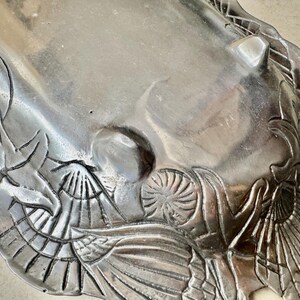May include: A silver-toned metal tray featuring an embossed design of various seashells and starfish along the edges. The tray has a smooth, reflective surface with raised decorative elements and small feet on the underside for stability.