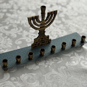 May include: A brass menorah with nine candle holders, set on a blue-grey base. The central holder is taller than the others. The menorah is placed on a white, patterned surface.