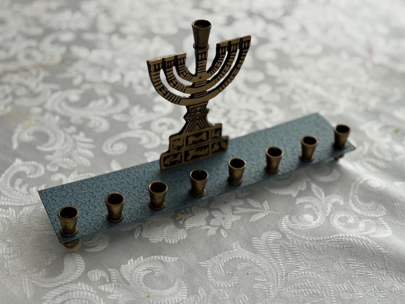 May include: A brass menorah with nine candle holders, set on a blue-grey base. The central holder is taller than the others. The menorah is placed on a white, patterned surface.
