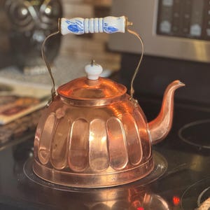 May include: A polished copper tea kettle with a rounded, fluted body and a curved spout. The kettle features a brass handle with a central white ceramic grip decorated with blue floral patterns, and a matching white ceramic knob on the lid.