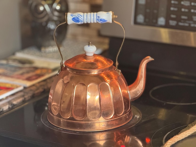 May include: A polished copper tea kettle with a rounded, fluted body and a curved spout. The kettle features a brass handle with a central white ceramic grip decorated with blue floral patterns, and a matching white ceramic knob on the lid.