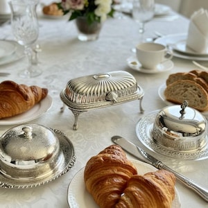 May include: A beautifully set table with a white tablecloth, featuring silver serving dishes, croissants, bread slices, and a cup of tea. The silver butter dishes have ornate designs, and the scene evokes a sense of elegance and fine dining.