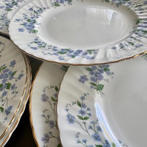 May include: Several white porcelain plates featuring a blue and green floral design. Each plate has a gold rim and a fluted edge. The plates are stacked, highlighting the detailed pattern and classic aesthetic, suitable for a formal dining setting.