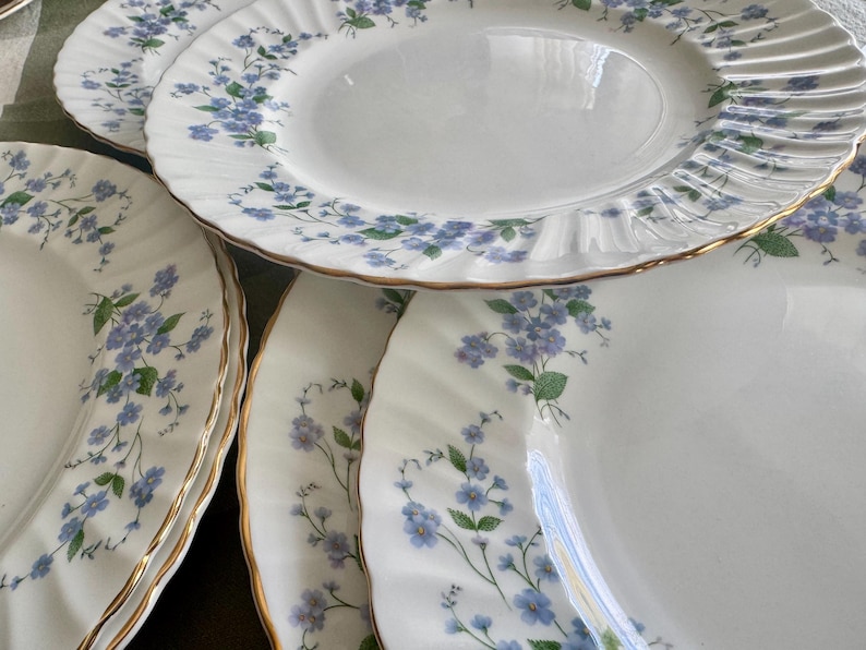 May include: A collection of white porcelain plates with a delicate floral pattern in blue and green. The plates have a gold trim and a scalloped edge. The plates are arranged in a stack, showcasing the intricate design and elegant style.