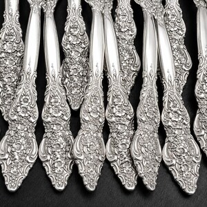 May include: A collection of silver-toned metal flatware handles arranged in a row. Each handle features a detailed, raised floral pattern with embossed blossoms and leaves covering the lower portion, transitioning into a smooth, polished stem.