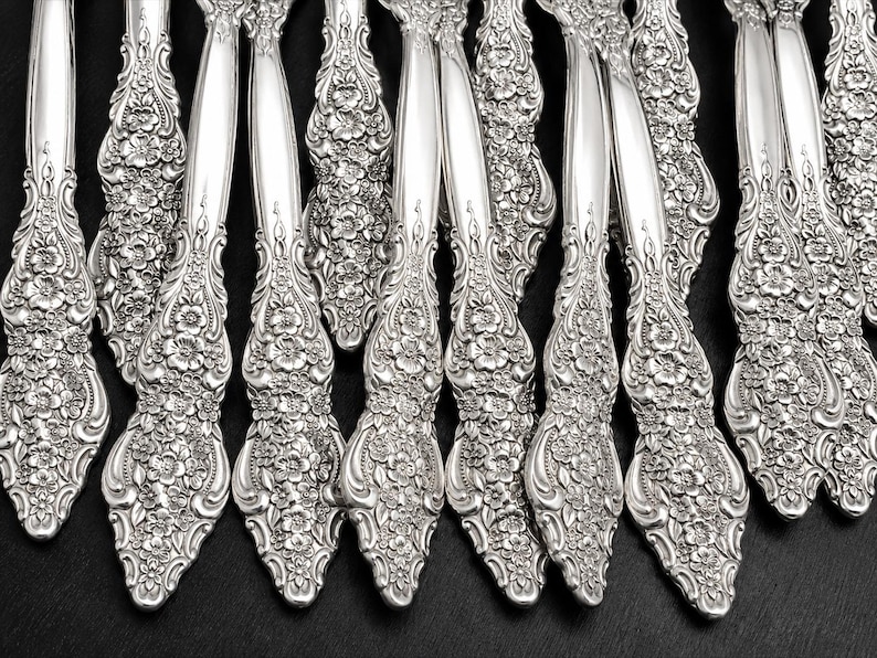 May include: A collection of silver-toned metal flatware handles arranged in a row. Each handle features a detailed, raised floral pattern with embossed blossoms and leaves covering the lower portion, transitioning into a smooth, polished stem.