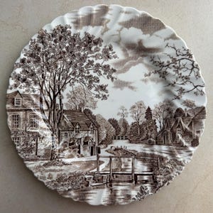 Pode incluir: Um prato redondo de cer&acirc;mica com borda recortada, apresentando uma estampa em tons de castanho e branco de uma paisagem rural. A cena retrata casas de pedra, uma &aacute;rvore grande, uma pequena ponte de madeira sobre um riacho e um c&eacute;u nublado.