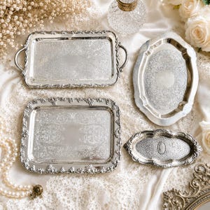 May include: An assortment of antique silver serving trays with ornate floral and scrollwork designs. The trays vary in shape, including rectangular and oval, and feature decorative edges and handles. The collection is arranged on a white lace surface.
