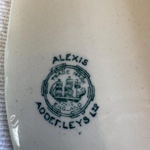 May include: A white ceramic surface featuring a dark green printed maker's mark. The mark includes the text ALEXIS, TRADE MARK, ENGLAND, and ADDERLEYS LTD, surrounding a central illustration of a sailing ship within a circular border.