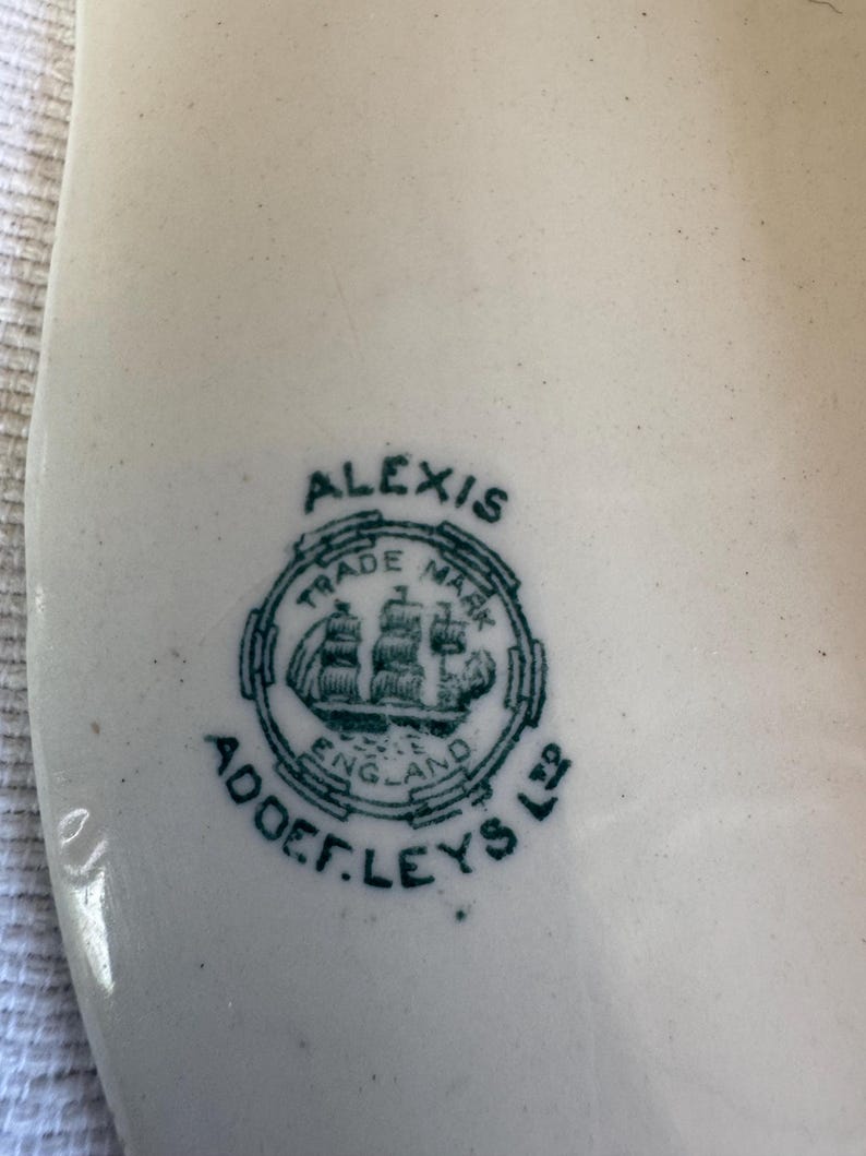 May include: A white ceramic surface featuring a dark green printed maker's mark. The mark includes the text ALEXIS, TRADE MARK, ENGLAND, and ADDERLEYS LTD, surrounding a central illustration of a sailing ship within a circular border.