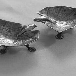 K&ouml;nnte beinhalten: Zwei antike Aschenbecher aus Silber. Jeder Aschenbecher hat die Form eines F&auml;chers mit aufwendigen Gravuren. Die Aschenbecher werden von Krallenf&uuml;&szlig;en getragen. Ein Aschenbecher hat eine eingebaute Zigarettenablage.