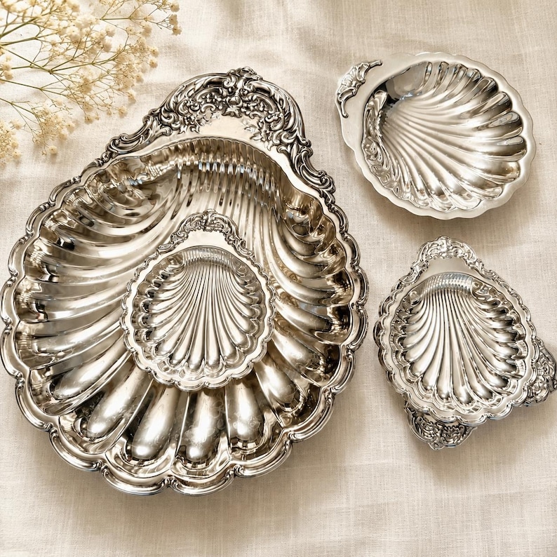 K&ouml;nnte beinhalten: Drei muschelf&ouml;rmige Schalen aus silberfarbenem Metall mit kunstvollen Pr&auml;gungen an den R&auml;ndern. Das Set besteht aus einer gro&szlig;en, einer mittleren und einer kleinen Schale, alle mit polierter, reflektierender Oberfl&auml;che und Rillen.