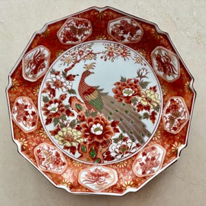 May include: An octagonal decorative plate featuring a central peacock illustration surrounded by red, white, and yellow floral patterns. The border displays a deep red background with gold scrollwork and hexagonal panels containing phoenix and butterfly motifs.