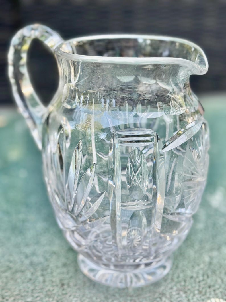 Puede incluir: Una jarra de cristal tallado transparente con asa y boquilla. La jarra presenta intrincados patrones geom&eacute;tricos y florales grabados en el cristal. El dise&ntilde;o incluye cortes verticales y angulados. La jarra se asienta sobre una base redonda.