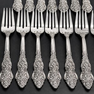 May include: A set of eight silver-toned metal forks arranged in two rows. Each fork features a four-tine head and a handle decorated with an intricate, embossed floral pattern that extends from the base of the tines to the rounded end.