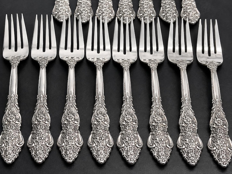 May include: A set of eight silver-toned metal forks arranged in two rows. Each fork features a four-tine head and a handle decorated with an intricate, embossed floral pattern that extends from the base of the tines to the rounded end.