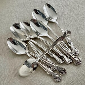 May include: A collection of silver spoons with ornate floral detailing on the handles. The spoons are arranged on a light gray surface, reflecting the surrounding environment. The spoons are likely for serving or eating.