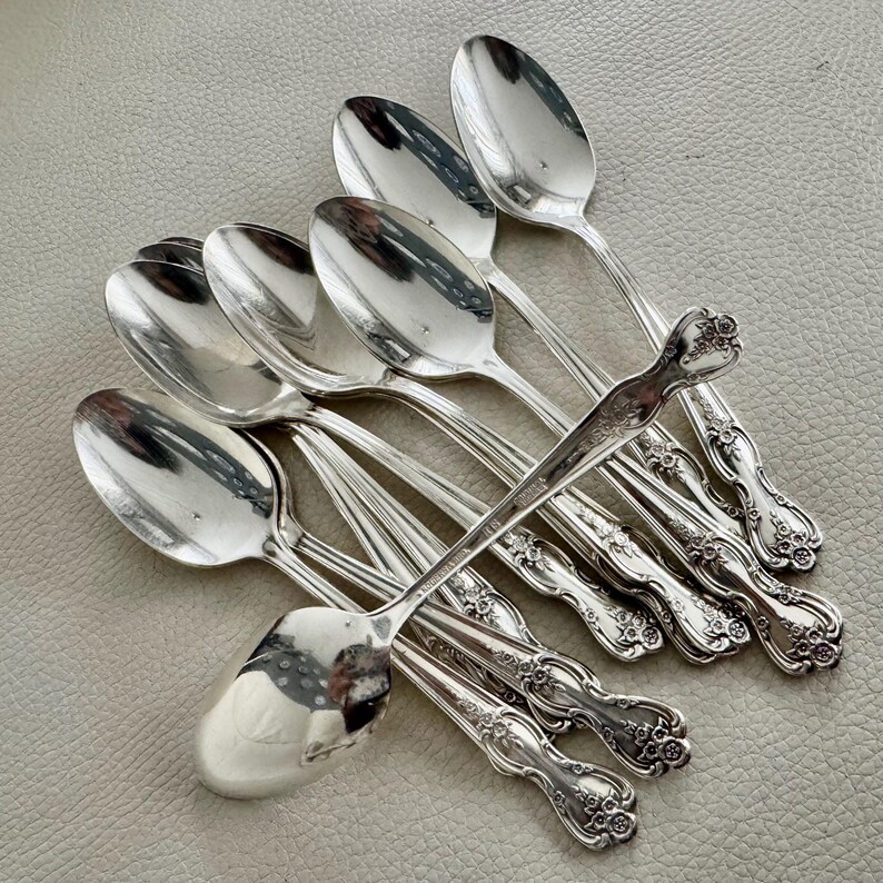 May include: A collection of silver spoons with ornate floral detailing on the handles. The spoons are arranged on a light gray surface, reflecting the surrounding environment. The spoons are likely for serving or eating.