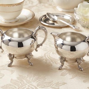 Puede incluir: Un azucarero de plata pulida con dos asas ornamentadas y tres patas decorativas, junto a una jarra de leche de plata a juego con pico vertedor, una asa y tres patas iguales, ambos con un acabado met&aacute;lico altamente reflectante.