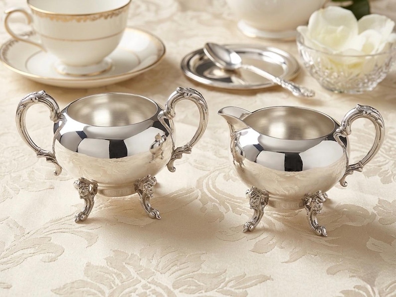 Puede incluir: Un azucarero de plata pulida con dos asas ornamentadas y tres patas decorativas, junto a una jarra de leche de plata a juego con pico vertedor, una asa y tres patas iguales, ambos con un acabado met&aacute;lico altamente reflectante.