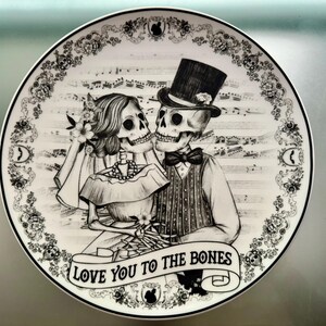 May include: A decorative plate featuring a black and white illustration of a skeleton couple in wedding attire. The plate has a floral border and musical notes in the background. The banner at the bottom reads "LOVE YOU TO THE BONES."