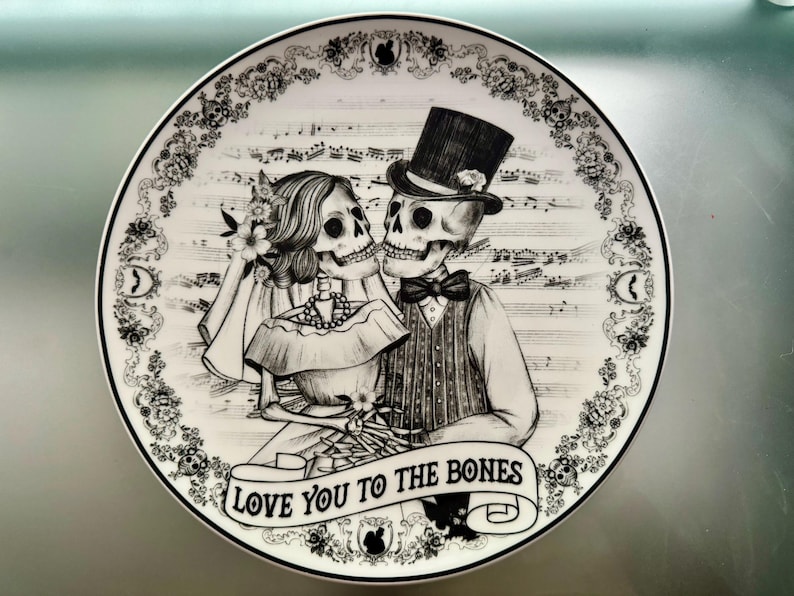 May include: A decorative plate featuring a black and white illustration of a skeleton couple in wedding attire. The plate has a floral border and musical notes in the background. The banner at the bottom reads "LOVE YOU TO THE BONES."