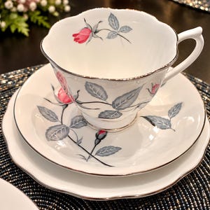 May include: A white porcelain teacup and saucer set featuring a delicate pattern of pink rosebuds and grey leaves. The edges of the cup and saucer are scalloped and finished with a thin silver trim, creating a refined and classic appearance.