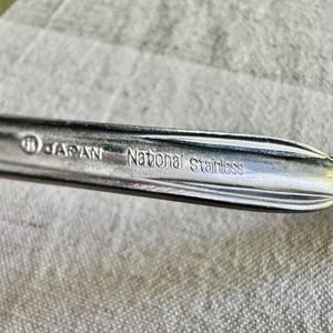 Może przedstawiać: Srebrzysty uchwyt sztućca ze stali nierdzewnej z wytłoczonym wzorem oraz wyraźnie wybitym napisem JAPAN i National stainless na powierzchni. Metal ma polerowane wykończenie z ozdobnymi żłobionymi liniami wzdłuż krawędzi.